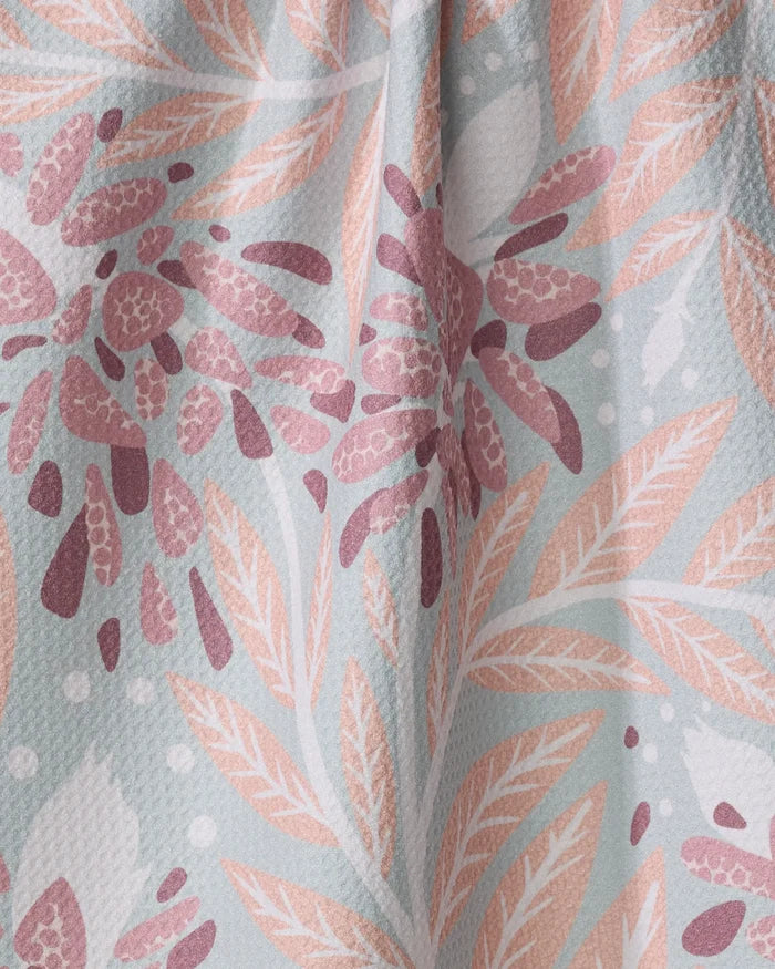 A close-up of the Bluebonnet tea towel by Julianne Haness, featuring a botanical pattern with peach and mauve leaves and flowers on light blue. The ultra-absorbent, woven texture blends style with functionality.