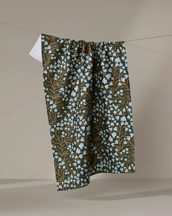The "Bluewood Garden" tea towel by Danelys Sidron features light blue flowers and brown branches on a dark fabric, hanging on a clothesline against a plain, light background.