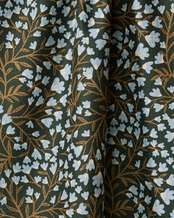 A close-up of Danelys Sidron’s Bluewood Garden tea towel shows small light blue flowers with brown stems on a dark green background. Made from recycled materials, it drapes softly, creating gentle folds and shadows.