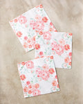 Three square "Blush Florals" napkins by Elena Fay, made from recycled materials, feature a pink and peach floral pattern with delicate green leaves, arranged elegantly on a light beige marble surface.