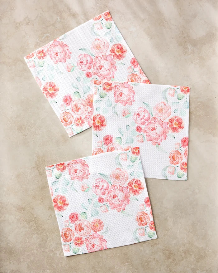 Three square "Blush Florals" napkins by Elena Fay, made from recycled materials, feature a pink and peach floral pattern with delicate green leaves, arranged elegantly on a light beige marble surface.