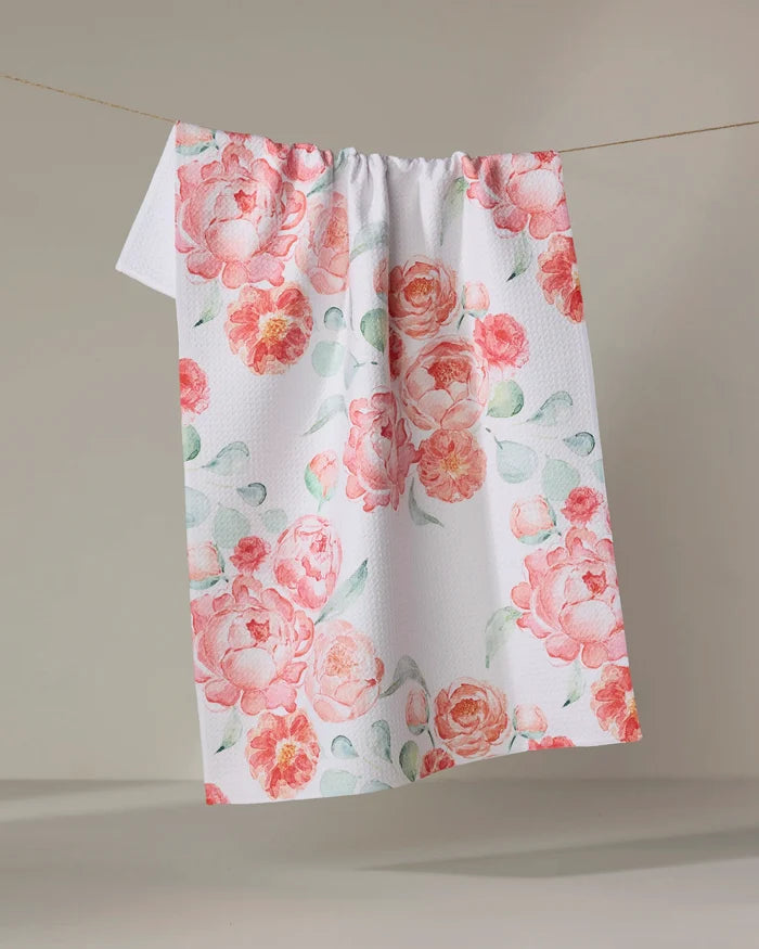 The "Blush Florals" kitchen towel by Elena Fay displays pink and peach watercolor-style flowers with green leaves on a white, textured fabric, hanging on a line against a neutral backdrop.