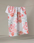 The "Blush Florals" kitchen towel by Elena Fay displays pink and peach watercolor-style flowers with green leaves on a white, textured fabric, hanging on a line against a neutral backdrop.
