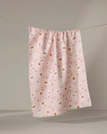The Sara Mari "Blush Meadows" tea towel, made from recycled materials and adorned with white and orange flowers, hangs on a clothesline against a neutral background.