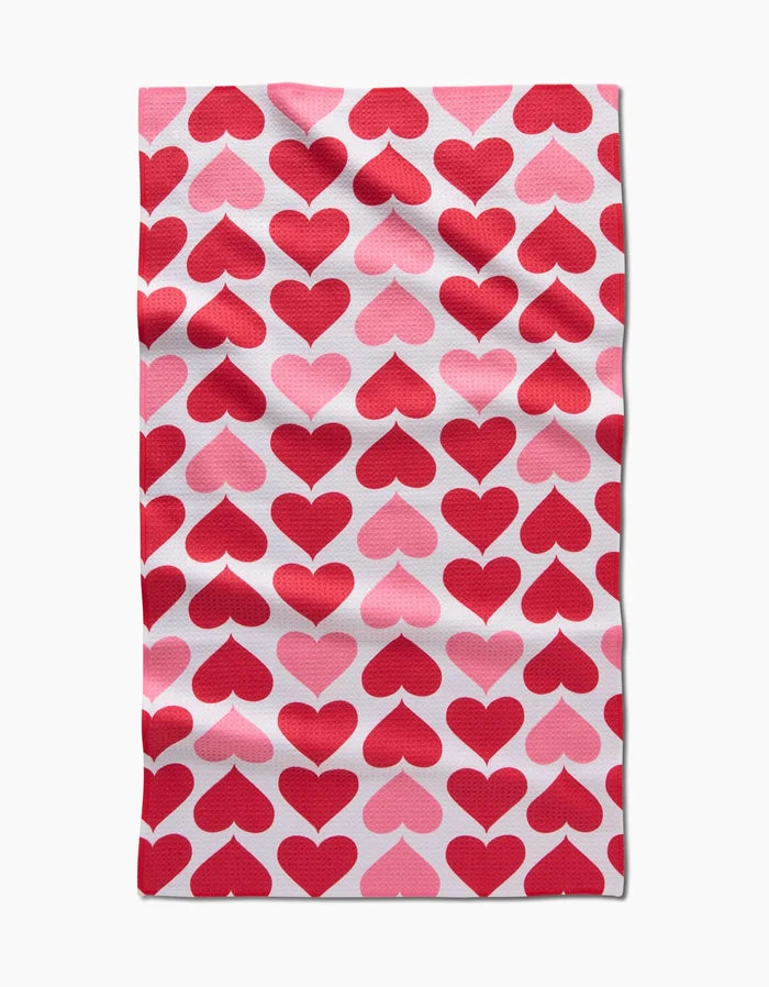 The Blushing Hearts tea towel by Danelys Sidron features rows of red and pink hearts on white, crafted from ultra-absorbent, recycled fabric with a naturally wrinkled look.