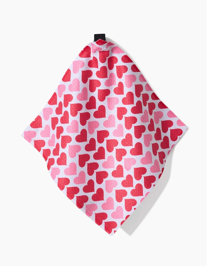 The Danelys Sidron Blushing Hearts is an absorbent dish towel featuring red and pink hearts on a light pink background, hanging from a black loop at the top.