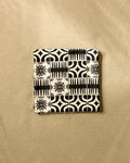 The GEOMETRY Bohemian Mosaic is a square recycled beach blanket featuring a black and white geometric and floral pattern, displayed on a textured beige surface.