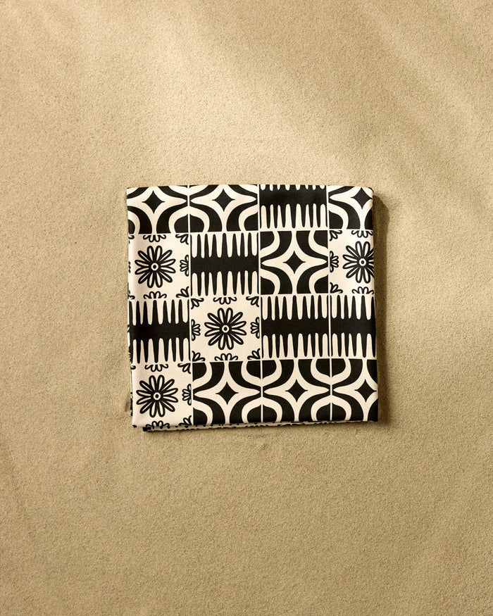 The GEOMETRY Bohemian Mosaic is a square recycled beach blanket featuring a black and white geometric and floral pattern, displayed on a textured beige surface.