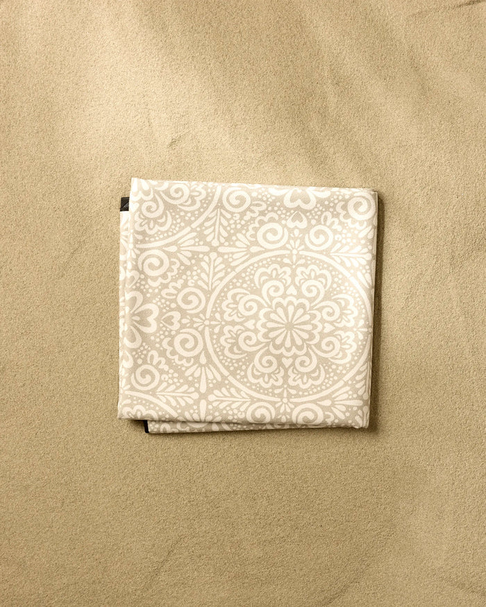 The Kris Ruff Boho Medallion recycled beach blanket, featuring a light color with an intricate white floral and geometric pattern, is shown folded on sand.