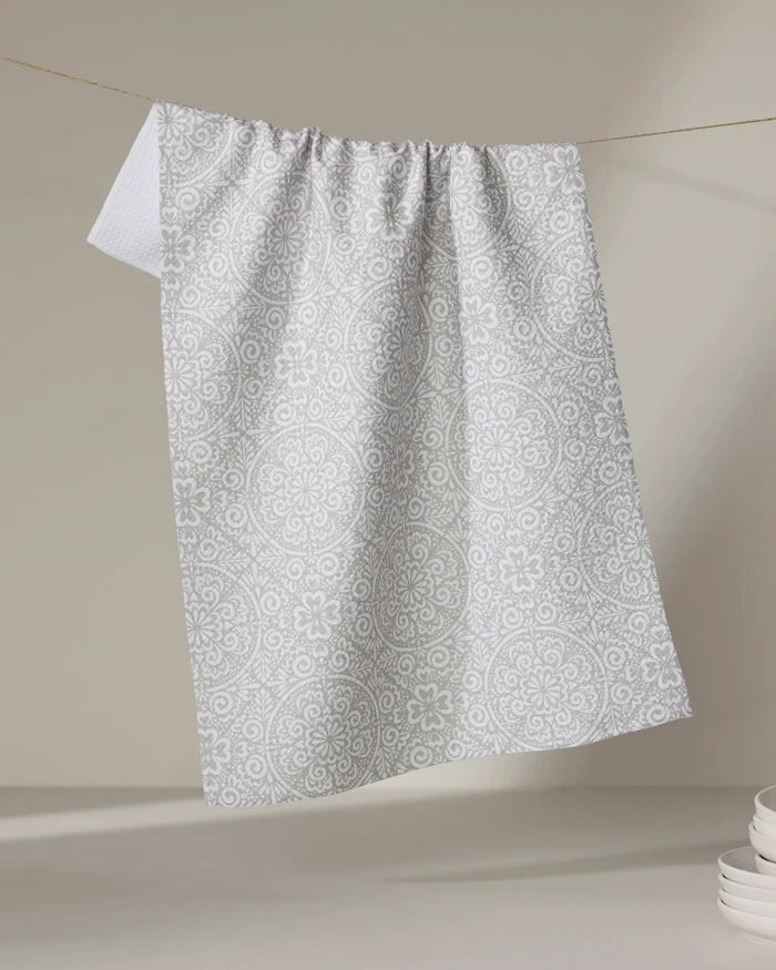 The Kris Ruff Boho Medallion dish towel, featuring a white and light grey pattern, hangs on a string against a neutral background, with a stack of white plates in the lower right corner.