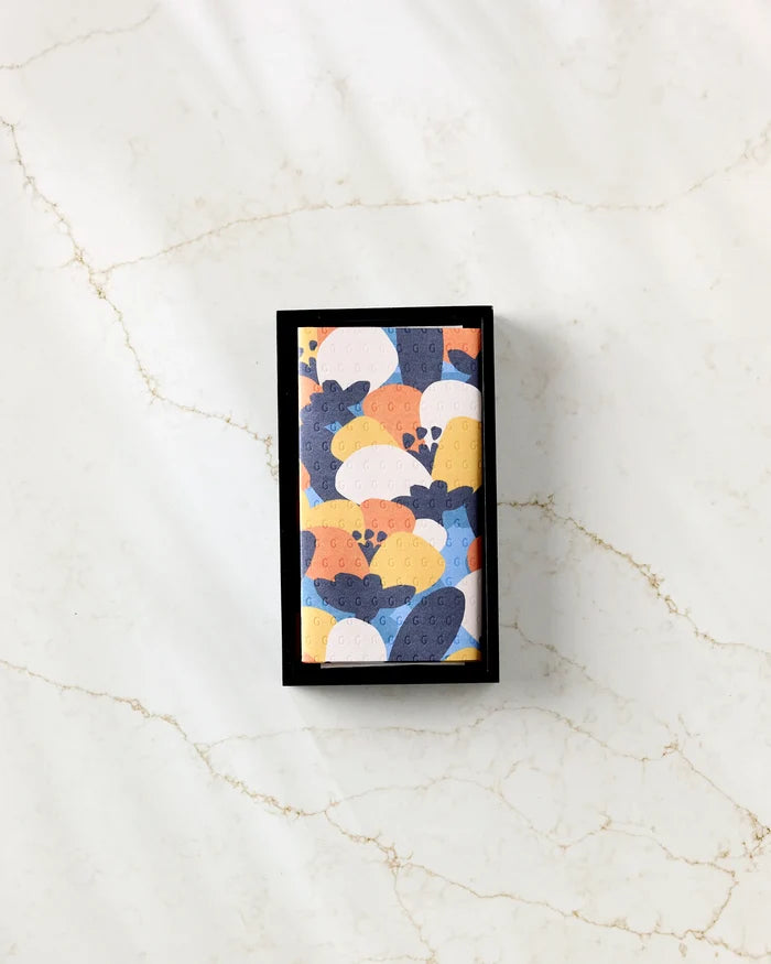 The Claire Iglesias Bold Petal Bloom eco-friendly kitchen towels, featuring a colorful abstract blue, yellow, white, and orange pattern, are displayed on a white marble surface with light brown veining.