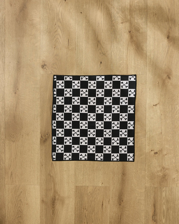 The Boneyard White Checkerboard towel by Charlie Rowan Designs features black and white checks with X and plus patterns, made from ultra-absorbent recycled materials, and is pictured on a light wooden floor.