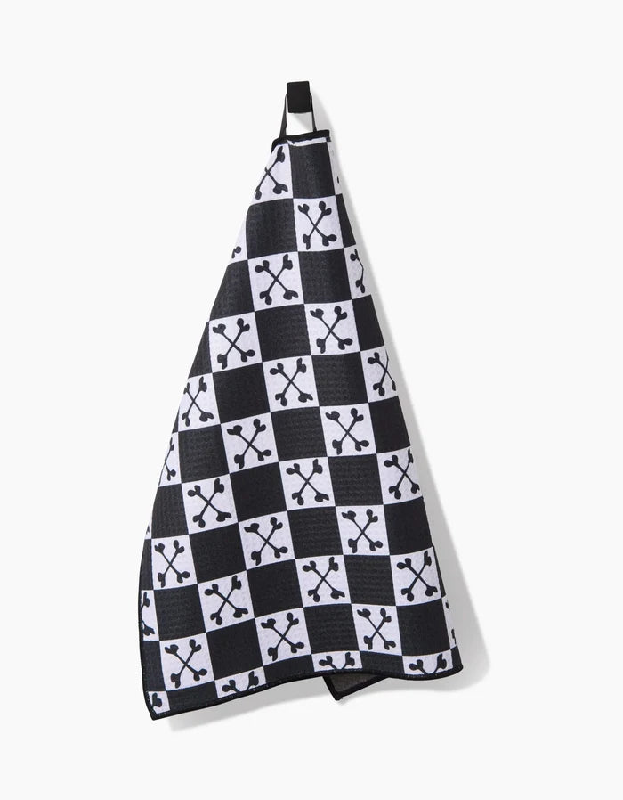 The Boneyard White Checkerboard Paw Towel by Charlie Rowan Designs hangs on a hook, showcasing a black and white checkered pattern with small crossed bones. Made from ultra-absorbent recycled materials, its stylish and eco-friendly.