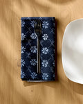 A fork rests on the Bonnie Indigo reusable napkin by Geometry, featuring a navy blue floral pattern, beside a white square plate on a light wooden table with shadows across the surface.
