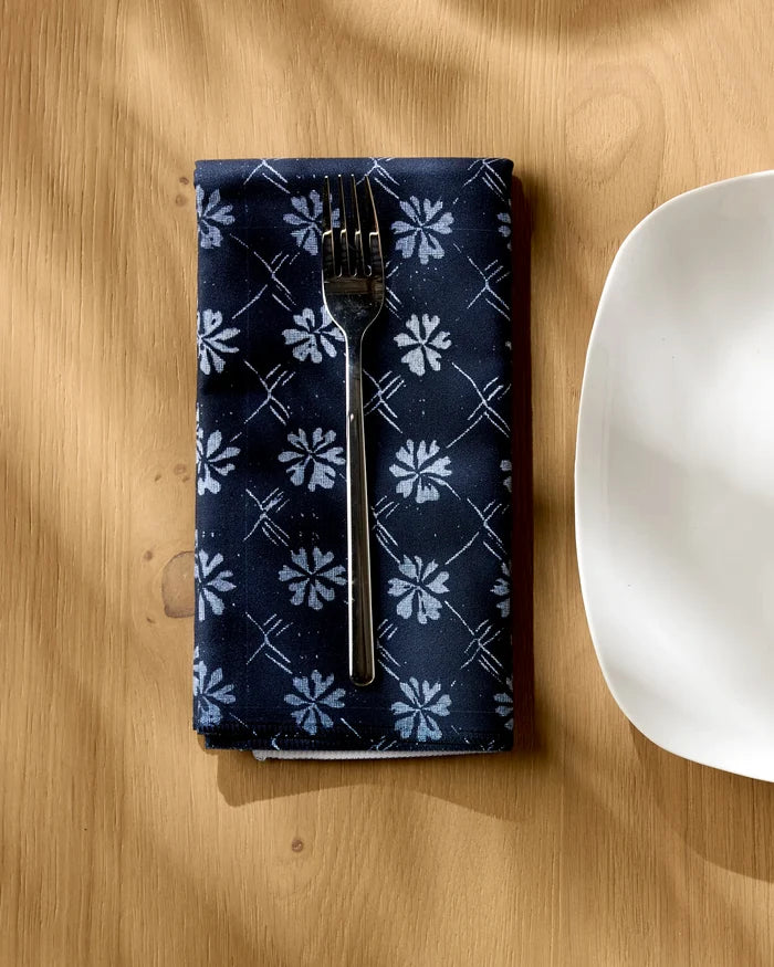 A fork rests on the Bonnie Indigo reusable napkin by Geometry, featuring a navy blue floral pattern, beside a white square plate on a light wooden table with shadows across the surface.