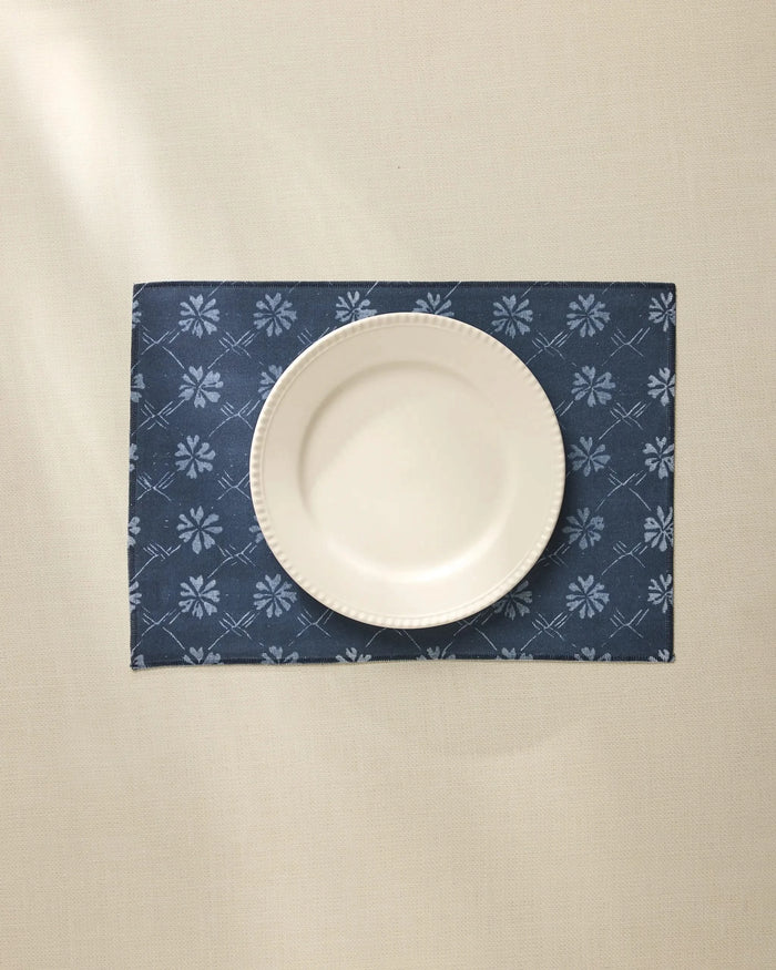 A white ceramic plate is centered on the Geometry Bonnie Indigo machine washable placemat, featuring a light floral pattern, atop a cream-colored tablecloth.