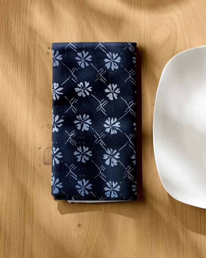 The Geometry Bonnie Indigo napkin, a folded navy blue reusable with white floral and grid patterns, rests on a light wooden table beside a white plate. Sunlight highlights this elegant recycled fabric accessory.