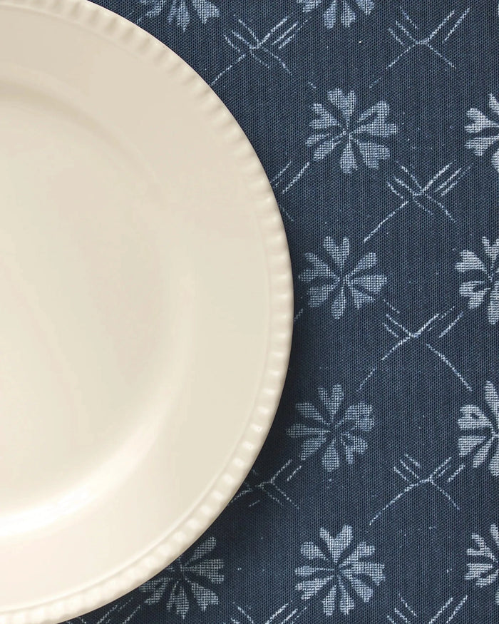A cream-colored plate with a scalloped edge sits on the left side of a dark blue Geometry Bonnie Indigo placemat, featuring white floral and geometric patterns.