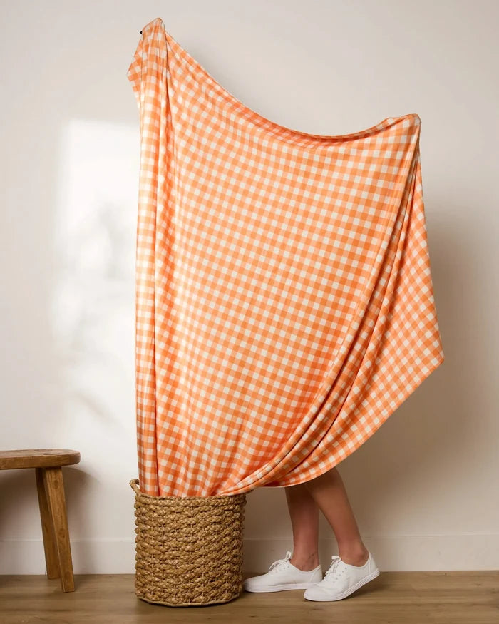 A person stands in a woven basket, mostly hidden behind the Geometry Boohoo Orange gingham throw draped over their head. Only their legs and white sneakers are visible, with a wooden stool nearby against a plain wall.