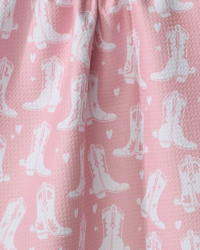 The "Boots On Worries Gone" kitchen towel by Aubrey Fairchild features pink textured fabric with white cowboy boots and hearts, softly gathered at the top—perfect for adding a playful, eco-friendly touch to your kitchen decor.