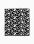 The Danelys Sidron Botanical Silhouettes Charcoal is a black square beach blanket made from quick-drying, recycled materials, showcasing a white floral pattern with evenly spaced clusters of flowers and leaves.