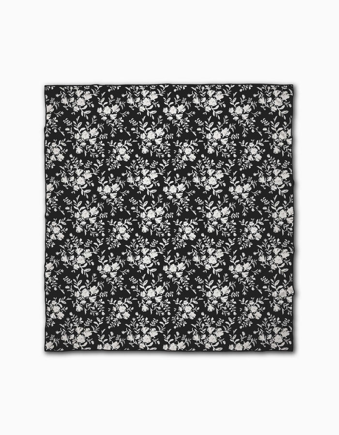 The Danelys Sidron Botanical Silhouettes Charcoal is a black square beach blanket made from quick-drying, recycled materials, showcasing a white floral pattern with evenly spaced clusters of flowers and leaves.