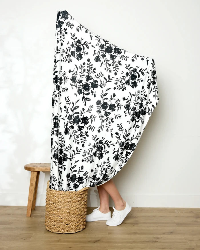 A person holds up the large white Danelys Sidron Botanical Silhouettes Charcoal blanket, featuring a black floral pattern. Only their lower legs and white sneakers are visible near a woven basket and wooden stool on a wood floor.