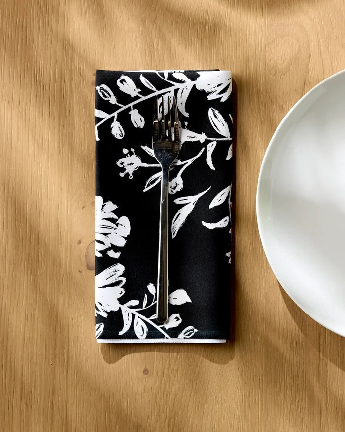 A silver fork rests on a Danelys Sidron Botanical Silhouettes Charcoal black recycled dinner napkin with white floral patterns, set on a wooden table next to a white plate, as sunlight casts soft shadows over the absorbent napkin.