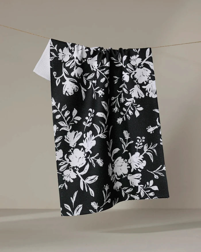 The Danelys Sidron Botanical Silhouettes Charcoal tea towel, featuring a white floral design on black and made from recycled materials, hangs on a thin string against a plain beige background.