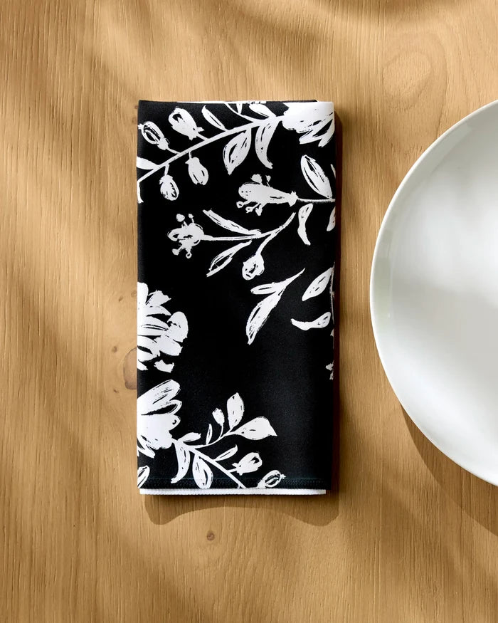 A Botanical Silhouettes Charcoal napkin by Danelys Sidron, with a white floral pattern, is neatly folded on a light wooden table beside a white plate, sunlight casting faint shadows on the scene.