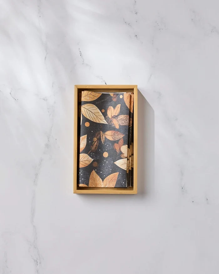 A wooden box of Geometry’s Botanical Wonderland cards, featuring gold leaves and dots on black, rests on white marble next to eco-friendly kitchen towels, all bathed in soft shadows.