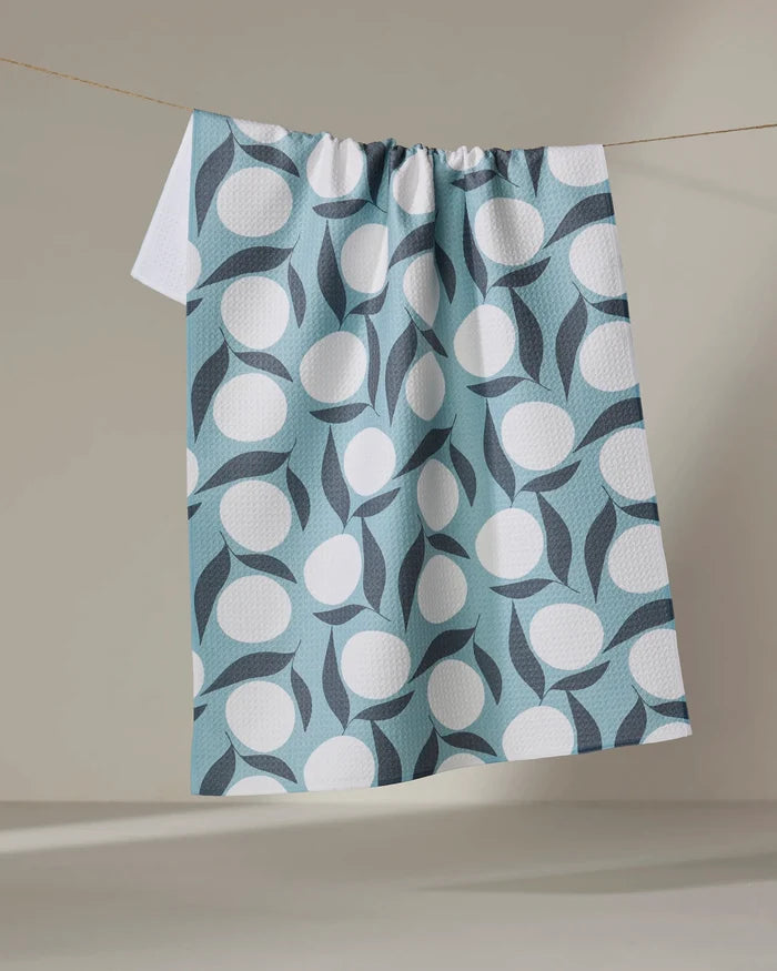The Bouncing Blooms towel by Lizzie Clark, featuring a geometric design with large white circles and dark blue curves on light blue, hangs on a line against a neutral wall.