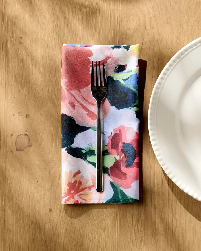 A silver fork lies on the "Bouquet" reusable dinner napkin by Ania Zwara Art, its colorful floral pattern beside a white scallop-edged plate on a light wooden table as sunlight casts soft shadows across the scene.
