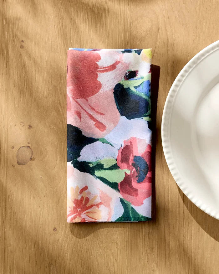 A folded "Bouquet" floral napkin from Ania Zwara Art rests on a light wood table by a white scalloped plate, with sunlight casting gentle shadows.