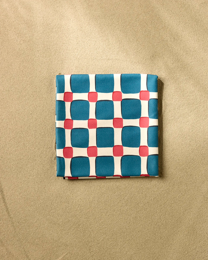 The Box Check Picnic by Hufton Studio is a micro-loop beach blanket featuring blue squares and red circles on white, neatly folded on a light brown textured surface.