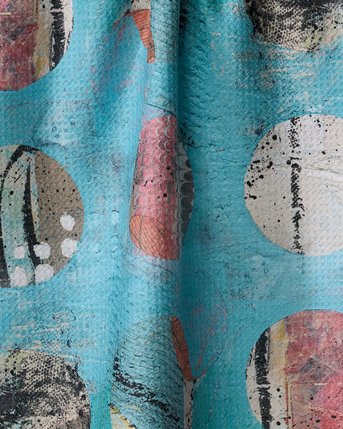 A close-up of "Boxed In" by Katherine Berkeley shows textured turquoise fabric with abstract circular patterns in white, black, pink, and beige. Made from recycled materials, it features speckled and striped designs and a slightly rough surface.