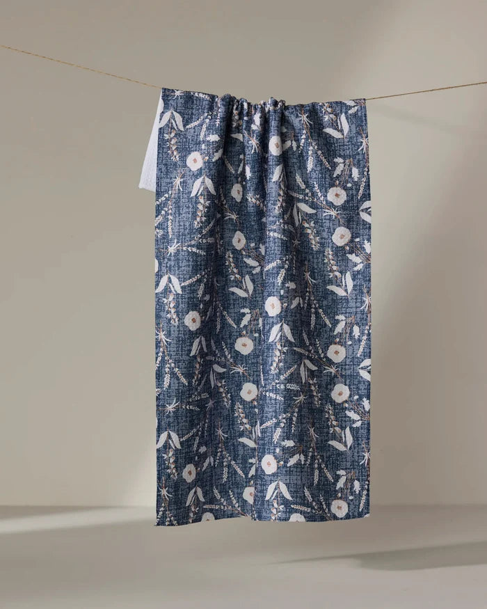 The Boyce Blue kitchen towel by Becki Owens features a blue and white floral pattern with subtle pink accents, hanging from a thin string against a plain, light-colored background.