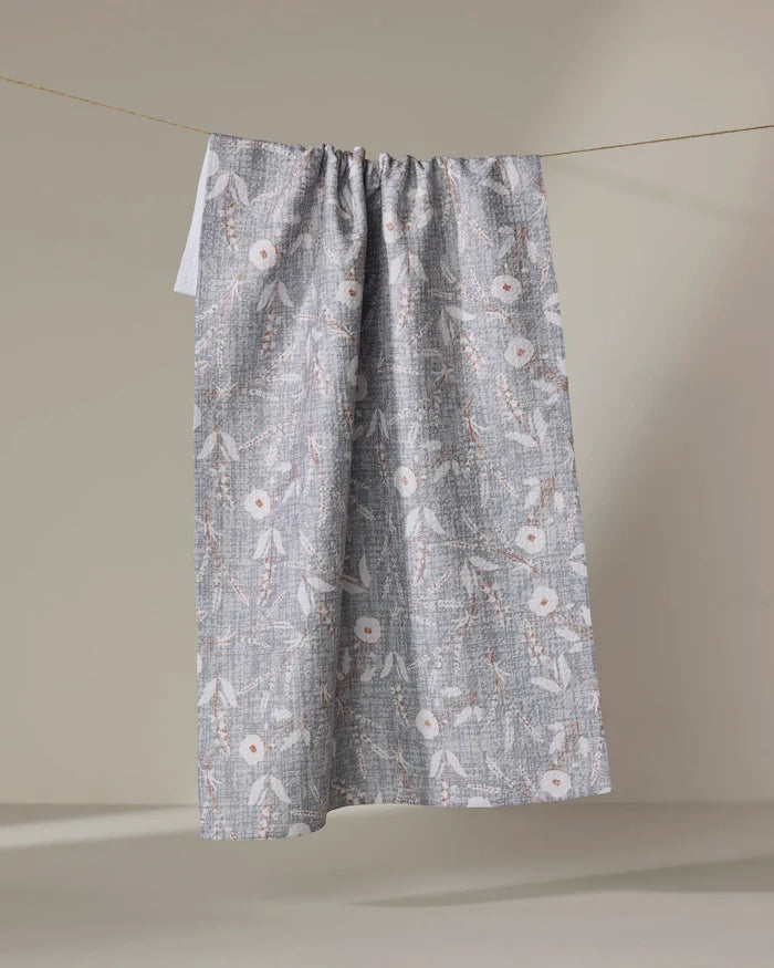 The Boyce Grey tea towel by Becki Owens, made from recycled materials and showcasing a white and beige floral pattern, hangs on a clothesline against a plain beige background.
