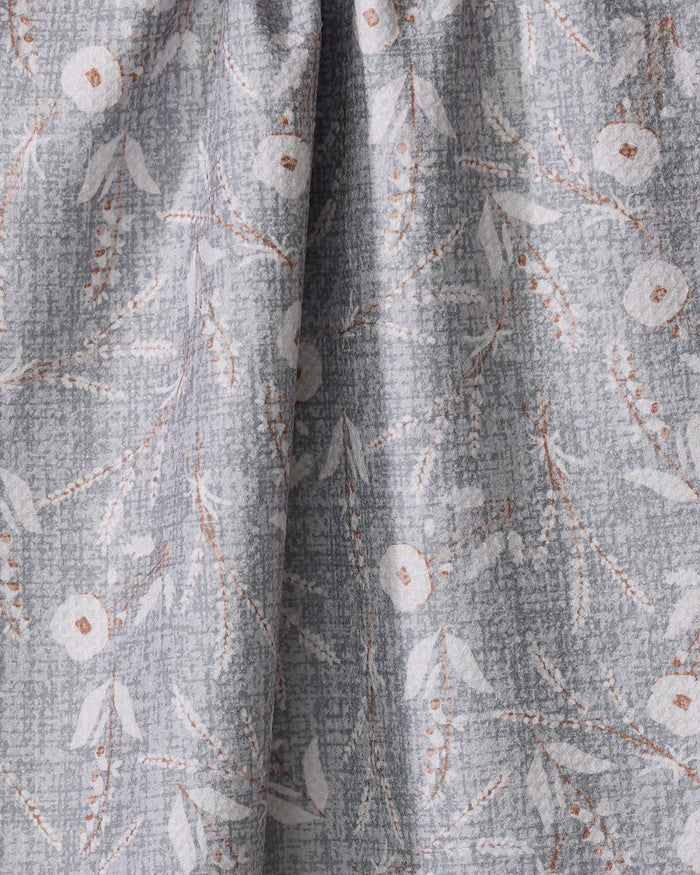 A close-up of the Boyce Grey kitchen towel by Becki Owens, crafted from recycled materials in light gray with a subtle floral pattern of white flowers and beige branches. The softly textured fabric has a gentle, elegant drape.