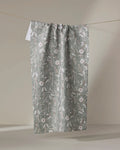 The Boyce Sage tea towel by Becki Owens, crafted from recycled materials, dries quickly and displays a white floral and leaf pattern. It is shown hanging on a string against a neutral beige background.