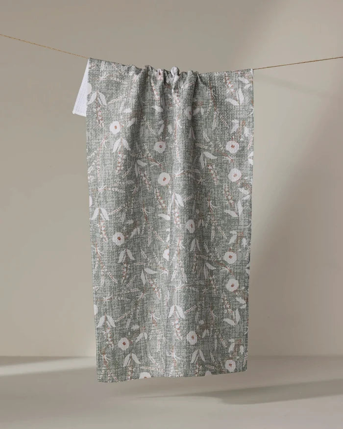 The Boyce Sage tea towel by Becki Owens, crafted from recycled materials, dries quickly and displays a white floral and leaf pattern. It is shown hanging on a string against a neutral beige background.