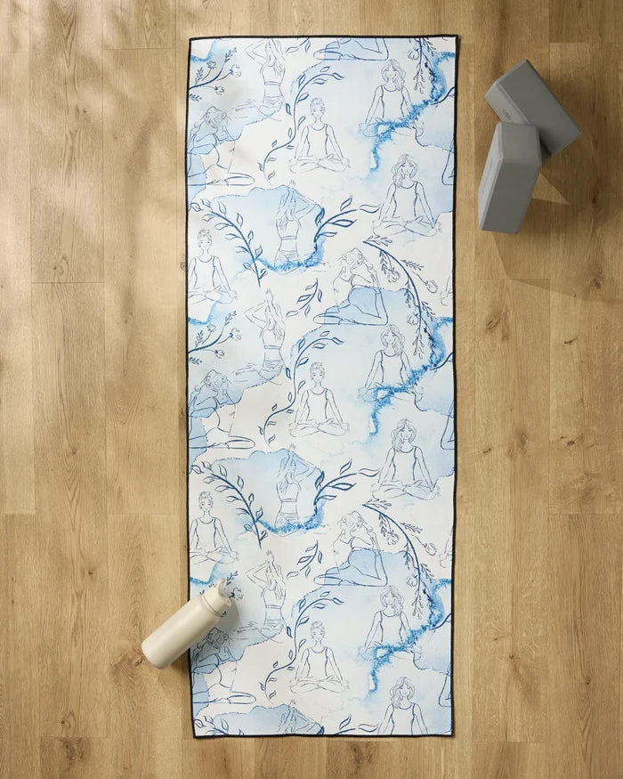 The "Breathe In Breathe Out" yoga mat by Elena Fay, featuring blue sketches of women in yoga poses and leafy branches, rests on a wooden floor beside a white water bottle, two gray blocks, and an eco-friendly yoga towel.