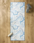 The "Breathe In Breathe Out" yoga mat by Elena Fay, featuring blue sketches of women in yoga poses and leafy branches, rests on a wooden floor beside a white water bottle, two gray blocks, and an eco-friendly yoga towel.