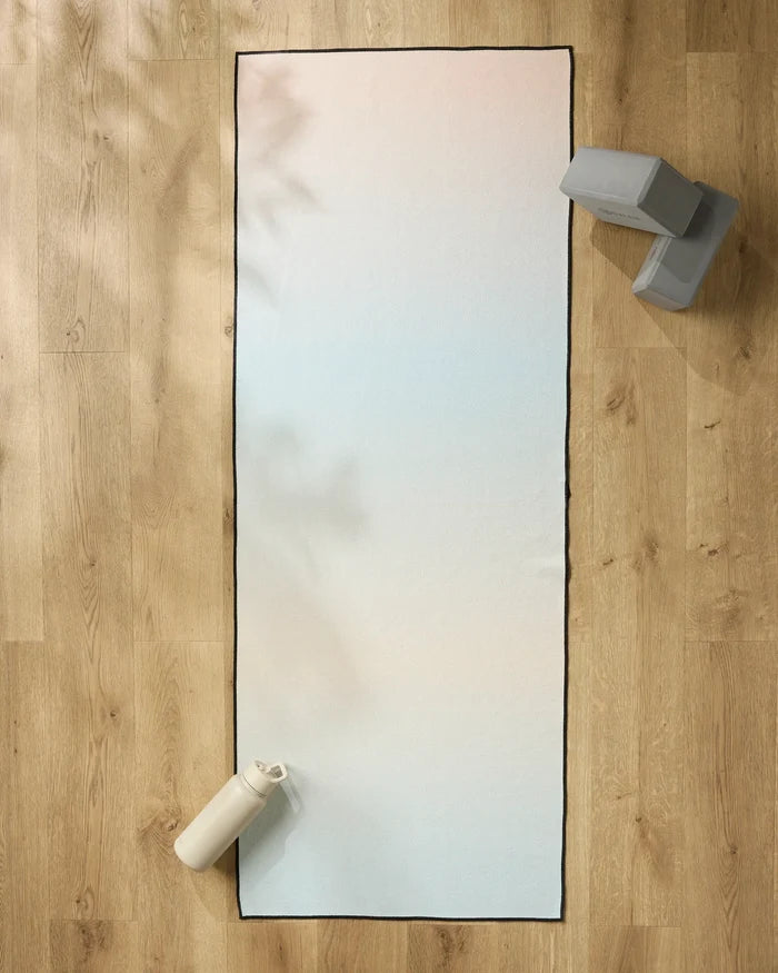 The GEOMETRY Breathe yoga mat lies on a wooden floor, with a water bottle at one end and a non-slip towel draped beside two grey yoga blocks at the other.