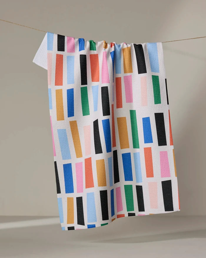 The Bricks tea towel by Jennifer Bouron features a colorful geometric pattern of vertical rectangles in blue, pink, orange, green, and black. Made from recycled materials, it dries quickly and adds sustainable style to any kitchen.