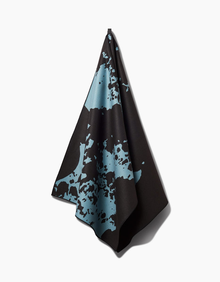 The Cole Strem Brush Stroke yoga towel, crafted from recycled materials, features bold black fabric with large, irregular light blue splatter and hangs from a single point against a plain white background.