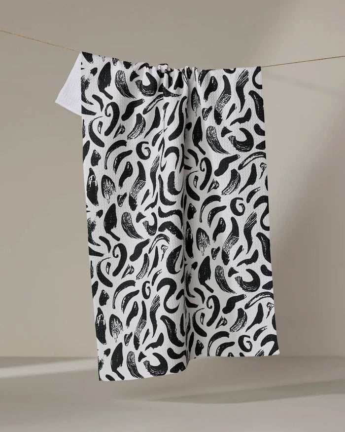 The Brush Strokes Swirls tea towel by Sarah Daugherty, crafted from recycled materials, features a bold black abstract pattern and hangs on a thin clothesline against a neutral background.