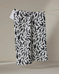 The Brush Strokes Swirls tea towel by Sarah Daugherty, crafted from recycled materials, features a bold black abstract pattern and hangs on a thin clothesline against a neutral background.