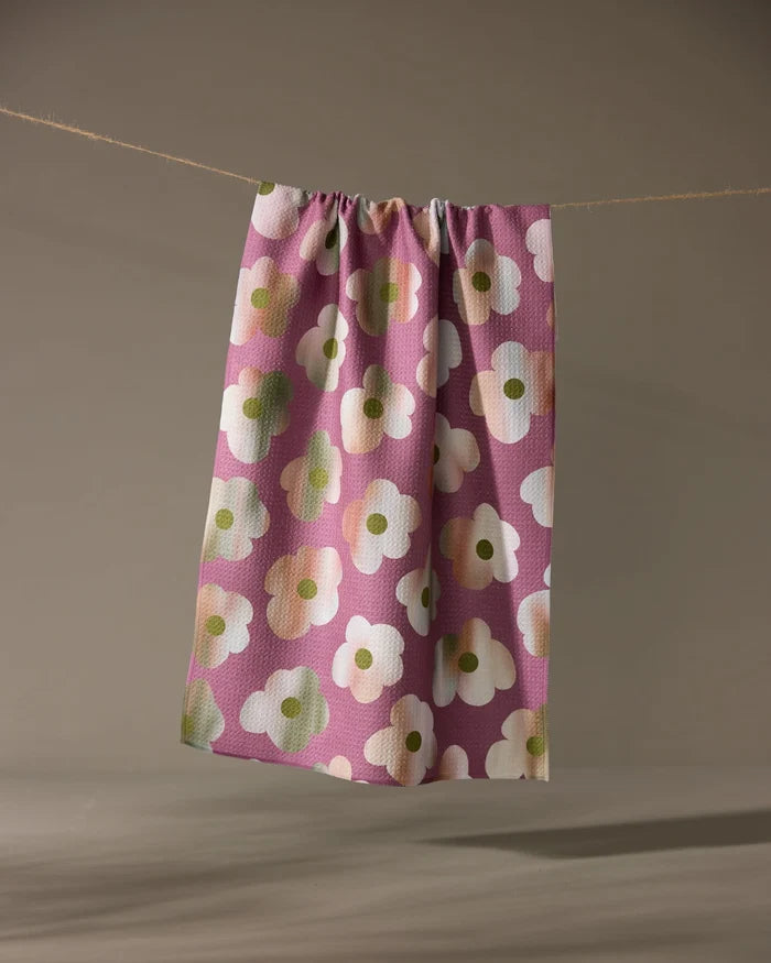 The Brushed Bouquet tea towel by Geometry, featuring bold white and green florals on pink and made from recycled materials, hangs from a clothesline against a neutral background.