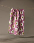 The Brushed Bouquet tea towel by Geometry, featuring bold white and green florals on pink and made from recycled materials, hangs from a clothesline against a neutral background.